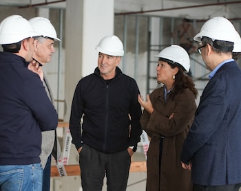 Cinco personas con cascos blancos de seguridad de pie en un sitio de construcción, con pilares de concreto y estructuras metálicas al fondo, discutiendo