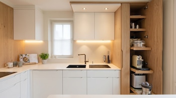 Una cocina moderna y luminosa con gabinetes blancos y paneles de madera clara, un fregadero negro, grifo metálico, una ventana y un armario con electrodomésticos.