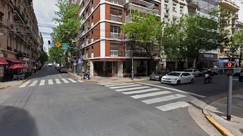 Calle de la ciudad con edificios de ladrillo y blancos, vehículos estacionados y en movimiento, árboles verdes, semáforos y un cruce peatonal marcado