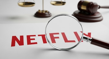 Primer plano del logo rojo de Netflix en papel blanco, con una lupa de borde metálico sobre él. Al fondo, borrosos, una balanza de la justicia y un mazo judicial.