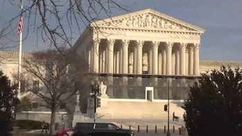 Vista frontal del edificio de la Corte Suprema de Estados Unidos con columnas clásicas, un cielo azul claro, un mástil con bandera y ramas de árboles en primer plano