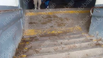 Vista cenital de una escalera de concreto gris con pasamanos metálicos y bandas antideslizantes amarillas, mostrando manchas de humedad y suciedad en los escalones