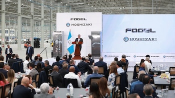 Hombre hablando en un podio junto a bandera de Guatemala. Pantallas con logos Fogel Hoshizaki, refrigeradores comerciales. Audiencia sentada en mesas. Nave industrial de fondo con montacargas