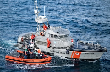 Un helicóptero blanco y naranja de la Guardia Costera y un efectivo con uniforme y casco sobre una pista de aterrizaje