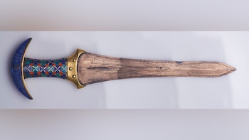 Daga antigua con empuñadura azul y con diseño de rombos verdes, rojos y dorados, con hoja de metal cobrizo desgastado sobre un fondo blanco