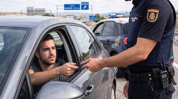 Un conductor entrega un teléfono móvil a un agente de policía uniformado en la ventanilla de un automóvil junto a una carretera.