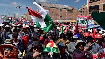 Multitud de personas, incluyendo maestros, protestando al aire libre bajo un cielo azul con nubes. Varias sostienen banderas de colores rojo, verde y blanco