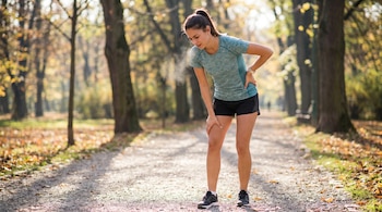 Mujer joven con camiseta verde y shorts negros en camino de tierra con árboles y hojas secas, sujetándose la espalda y la pierna, con vaho en la boca.
