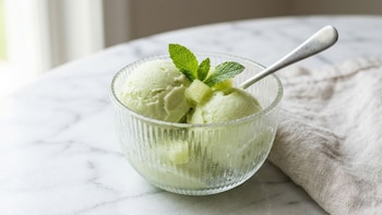 Cuenco de vidrio estriado con dos porciones de helado verde claro, hojas de menta y una cuchara metálica sobre una mesa de mármol