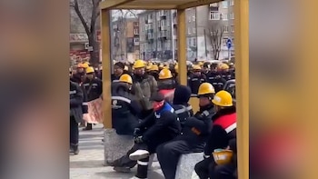 Multitud de trabajadores con cascos amarillos y uniformes oscuros se congregan en una calle urbana de Rusia, con edificios residenciales al fondo