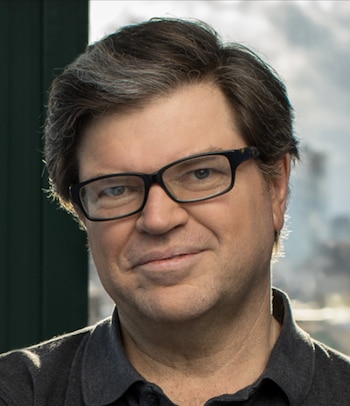 Retrato de Yann LeCun, un hombre con cabello oscuro y canoso, gafas de montura negra, y camisa polo gris, mirando y sonriendo ligeramente
