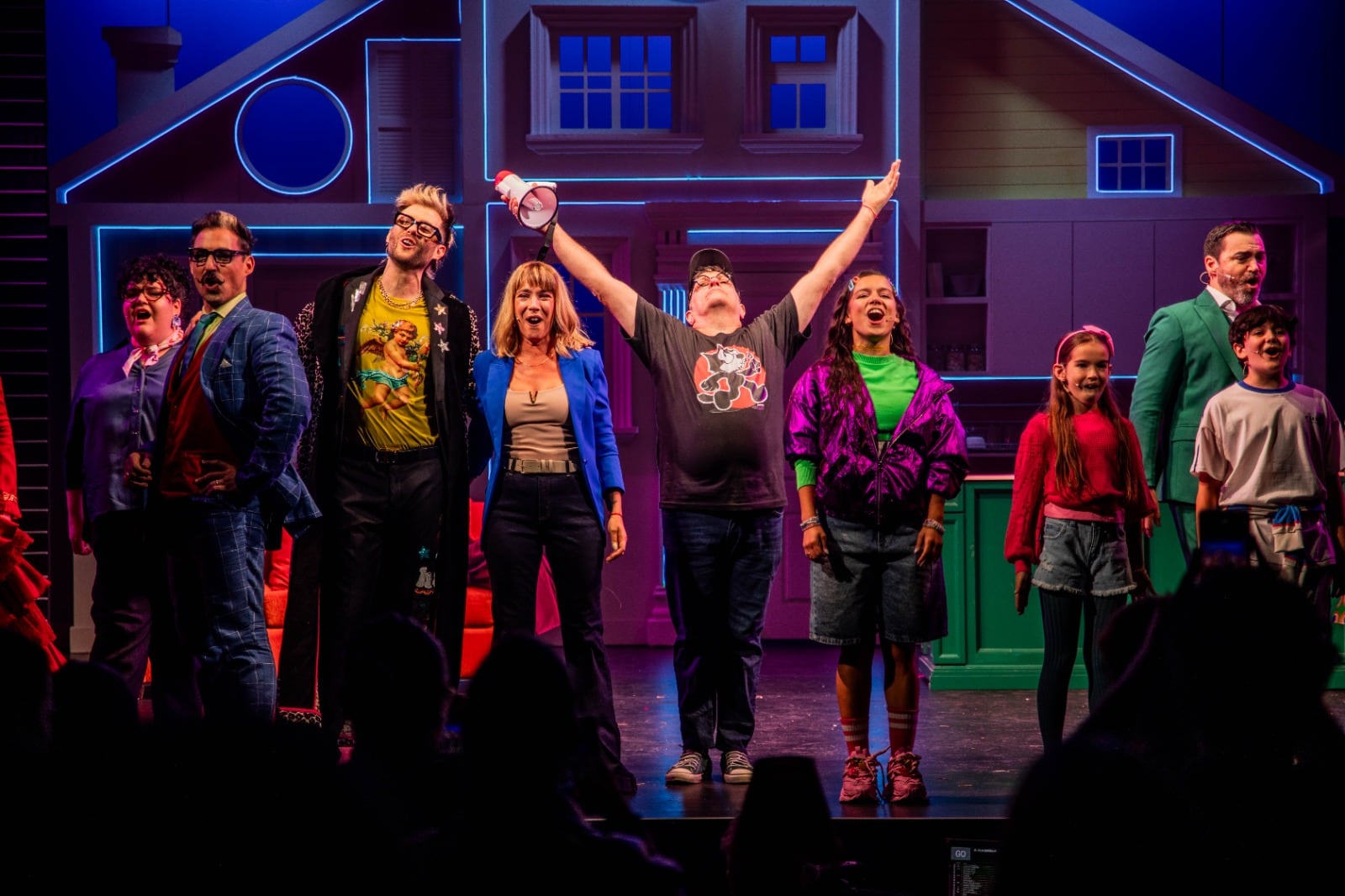 Los actores del musical 'Papá por siempre' saludan entusiasmados a la audiencia desde el escenario con un vibrante telón de fondo de una casa moderna tras finalizar una función.