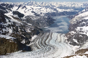 Vista aérea de un glaciar con vetas de sedimento desembocando en una bahía azul, flanqueado por montañas rocosas y nevadas bajo un cielo claro