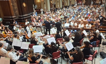 Músicos de una orquesta tocan instrumentos de cuerda y viento frente a una directora de orquesta y un público sentado en bancos de madera