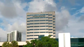 Edifício bege de 14 andares com janelas azuis e o nome 'UNIFORM ADVANTAGE' acima, visto sob um céu nublado. Árvores verdes na frente