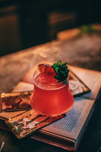 Un cóctel rojo intenso en un vaso de cristal reposa sobre un libro abierto y varias cartas de tarot, adornado con una rodaja de cítrico y hojas de menta