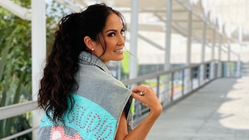 Joanna Vega-Biestro sonriendo y mirando por encima del hombro, con cabello oscuro recogido y un chaleco de mezclilla azul claro con adornos