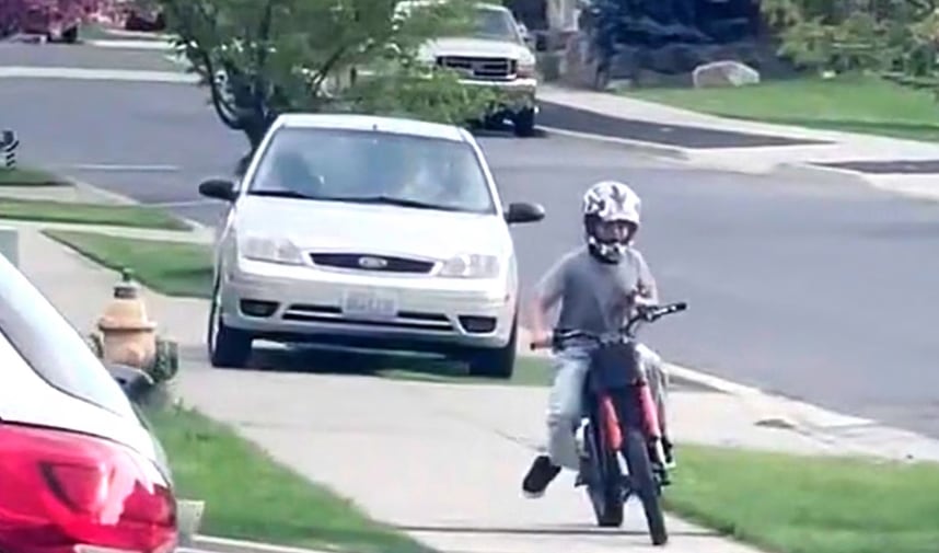 Un niño con casco conduce una minimoto por la acera, pasando junto a coches aparcados en una calle residencial (Cindy Hagan)