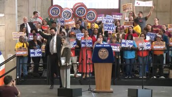 Multitud de personas con pancartas en una manifestación, con dos hombres en podios. Un podio tiene el sello de la ciudad de Nueva York. Las pancartas mencionan viviendas asequibles y responsabilidad de propietarios