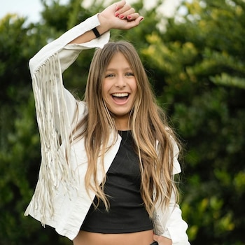Niña sonriente con cabello largo y castaño, viste chaqueta blanca de flecos y blusa negra, brazo derecho levantado, uñas pintadas, fondo verde difuminado