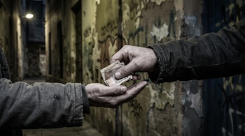 Primer plano de dos manos en un callejón oscuro. Una mano entrega billetes a la otra que tiene la palma extendida. Paredes con grafitis borrosos de fondo.