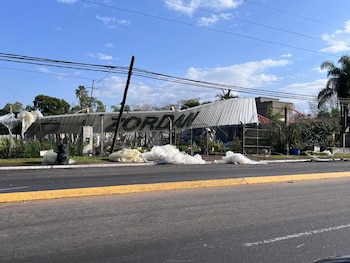 Edificio con techo metálico y poste de luz caídos, desechos blancos plásticos esparcidos, vegetación, carretera con línea amarilla, cielo azul