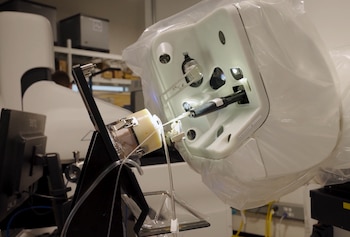 Máquina blanca de laboratorio con cubierta de plástico transparente, mostrando un brazo que interactúa con una muestra en un soporte con pinzas y tubos