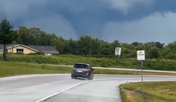 Un tornado oscuro en el cielo sobre una carretera mojada con un coche negro, casas y árboles al fondo. Una señal de No Estacionar