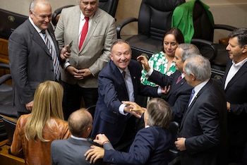 Grupo de políticos, mayormente hombres con traje, interactuando en un recinto oficial. Un hombre con traje azul sonríe mientras da la mano a otro