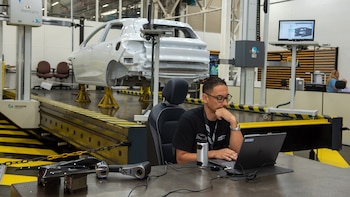 Un hombre con gafas y camiseta de Chevrolet trabaja en una laptop; detrás, un chasis de coche blanco en soportes y maquinaria de medición industrial