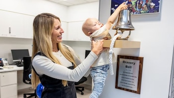 Una mujer rubia sonriente sostiene a un bebé que estira el brazo para tocar una campana de plata brillante montada en la pared con una placa