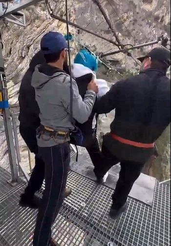 Tres personas sujetan a un participante con casco azul en una plataforma metálica junto a un acantilado rocoso