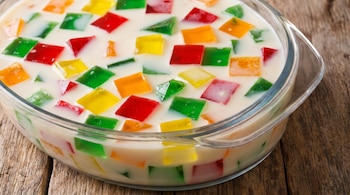 Tazón de vidrio transparente con gelatina mosaico, mostrando cubos de gelatina de colores rojo, verde, amarillo y naranja inmersos en una base blanca lechosa, sobre madera.