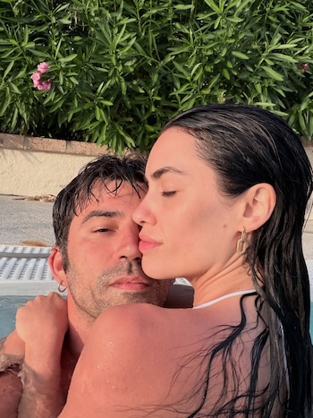 Una mujer y un hombre de cabello oscuro y mojado se abrazan en una piscina, con vegetación verde y algunas flores rosas de fondo