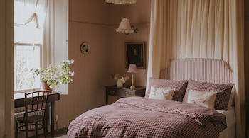Dormitorio con cama de patrón a cuadros, cabecero de rayas, dos almohadones blancos, mesa de noche, lámpara, cuadro, escritorio y silla de madera, planta junto a ventana.