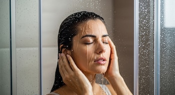 Primer plano de una mujer de cabello oscuro con ojos cerrados bajo el agua de la ducha. Gotas de agua caen por su rostro y cabello mojado, con sus manos a los lados de la cabeza. Cristales de ducha con gotas de agua visibles.