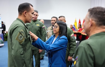 Una mujer con blazer azul coloca una insignia en la solapa de un hombre con uniforme militar verde. Otros militares y banderas venezolanas se ven de fondo