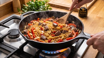 Una sartén de hierro con fideos, trozos de carne, morrón, cebolla, ajo y ají sobre una estufa con fuego. Una mano revuelve la comida con una cuchara de madera.