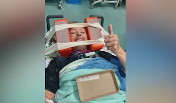 Una mujer permanece recostada en una camilla con la cabeza inmovilizada mediante bloques naranjas mientras levanta el pulgar
