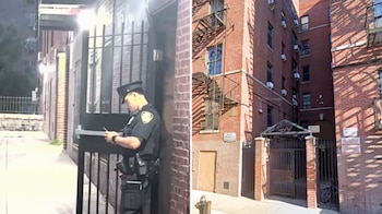 Policía uniformado mira un teléfono móvil junto a una verja negra. Al lado hay edificios de ladrillo con escaleras de incendio, ventanas, aires acondicionados y una boca de incendio