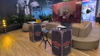 Dos estaciones de juego con pantallas que muestran el videojuego Spider-Man, consolas PlayStation 5 y mandos. Pedestales con diseño de telarañas. Sofás y plantas.