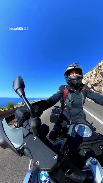 Vista en primera persona de una mujer con casco y chaqueta de moto conduciendo por una carretera costera. El mar azul se ve a la izquierda y montañas rocosas a la derecha