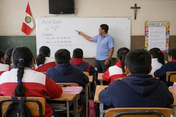 Un profesor señala una pizarra blanca a un grupo de alumnos sentados de espaldas en un aula. Hay una bandera de Perú, un crucifijo y un cartel de normas.