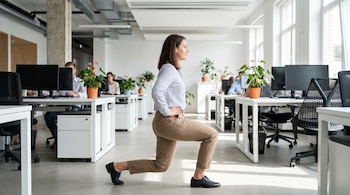 Una mujer de perfil hace una zancada inversa en una oficina con escritorios, ordenadores, sillas y plantas. Varias personas trabajan al fondo.