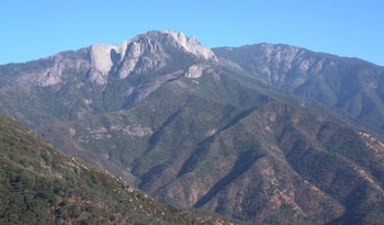 Vista panorámica de montañas con cumbres rocosas y laderas cubiertas de árboles, vegetación densa en los valles, y cielo azul despejado