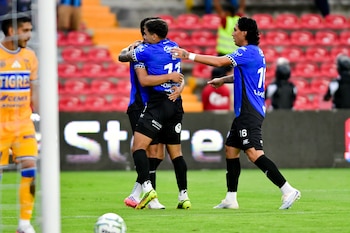 Tres futbolistas con uniformes azules y negros se abrazan, un cuarto futbolista con uniforme amarillo y azul se ve borroso, un balón de fútbol, asientos rojos de estadio