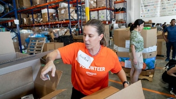 Mujer con camiseta naranja "Amazon Volunteer" y placa "Adriana" empaca una caja en un almacén con estantes llenos de paquetes y otras personas