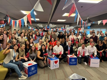Numeroso grupo de personas sonriendo, muchos con objetos rojos en las manos, en un interior decorado con guirnaldas y globos, junto a cajas con el logo de Philip Morris Argentina