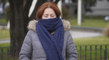 Mujer de cabello castaño rojizo con bufanda azul marino que cubre boca y nariz, campera gris con capucha de piel. Fondo borroso de árboles y valla.