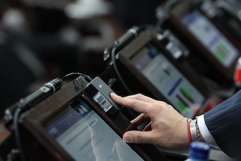 Primer plano de una mano presionando un escáner de huellas dactilares sobre un dispositivo electrónico en un atril, con pantallas borrosas de fondo
